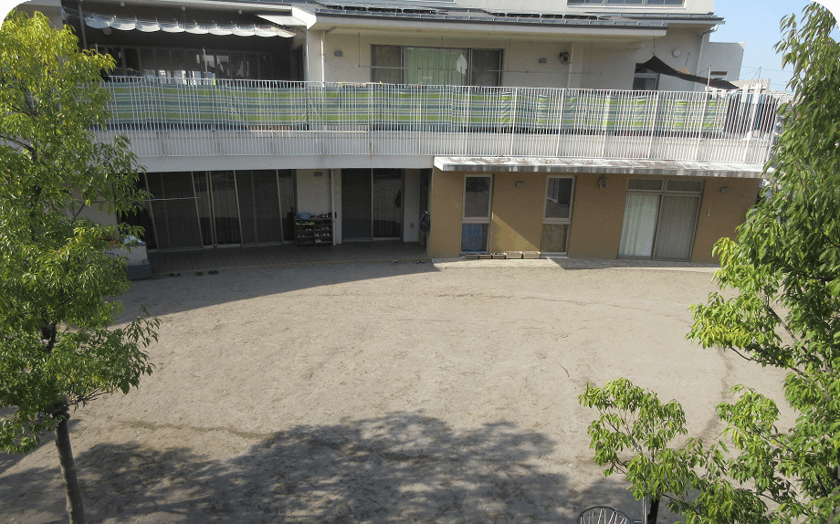 日差しが降り注ぐ園庭と、2階建ての建物がある保育園の風景